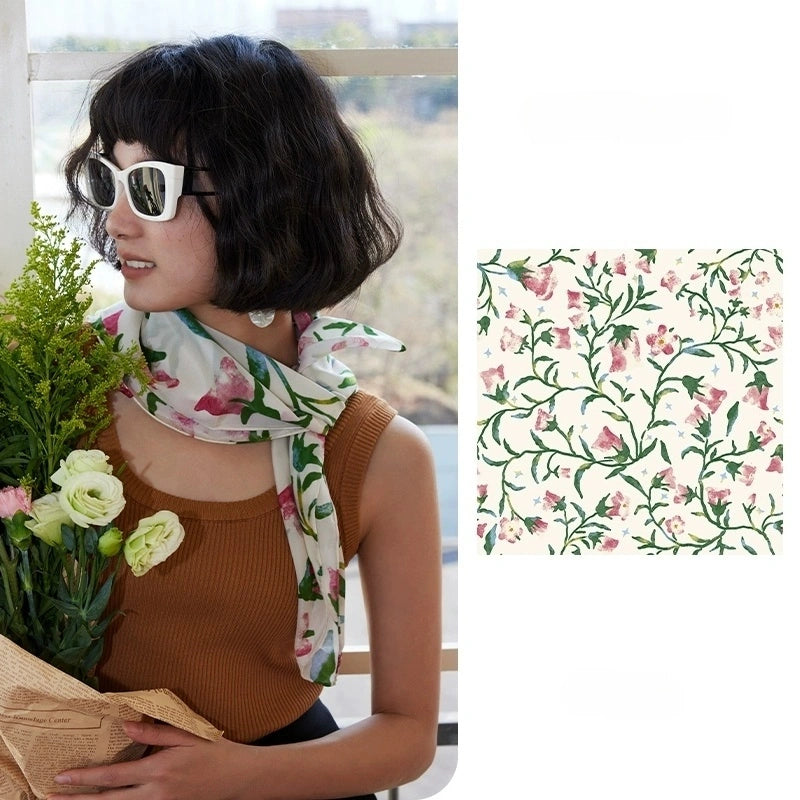 Woman wearing a floral silk square scarf with pink morning glory print as a neck accessory, holding a bouquet of white flowers against a window backdrop.