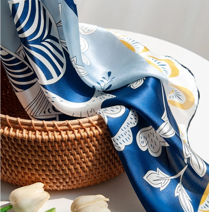 Blue floral and bird print silk scarf styled on a woven basket for elegant accessory display.