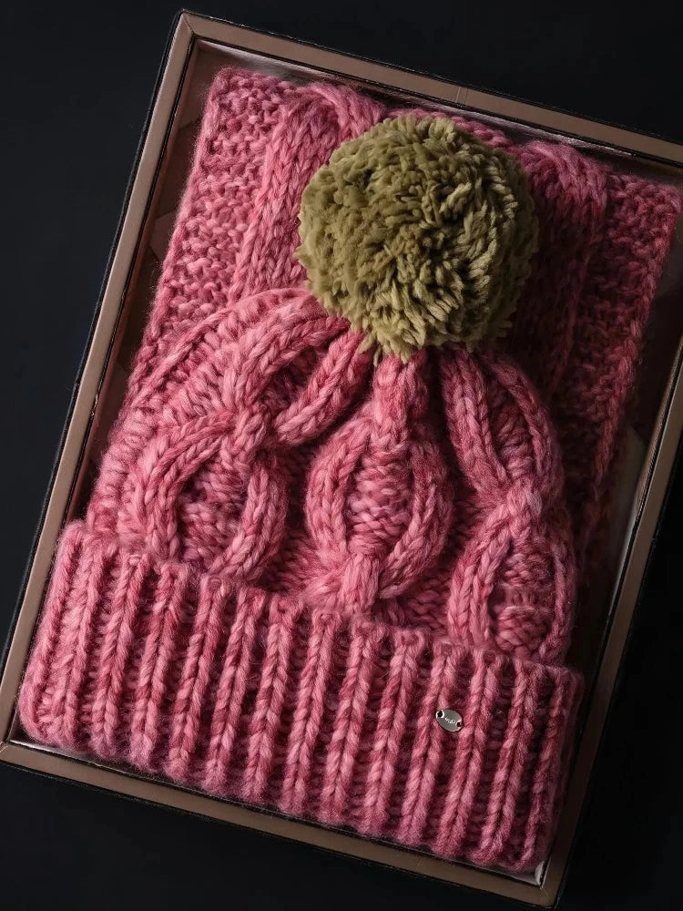 Dusty pink cable knit wool beanie with an olive-brown pom pom and ribbed cuff, displayed in a box.