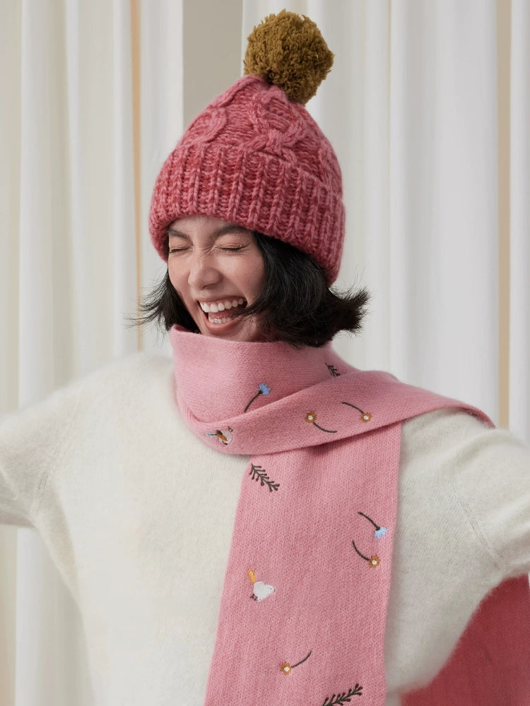 Model wearing a dusty pink cable knit beanie with a contrasting brown pom pom and pink scarf.