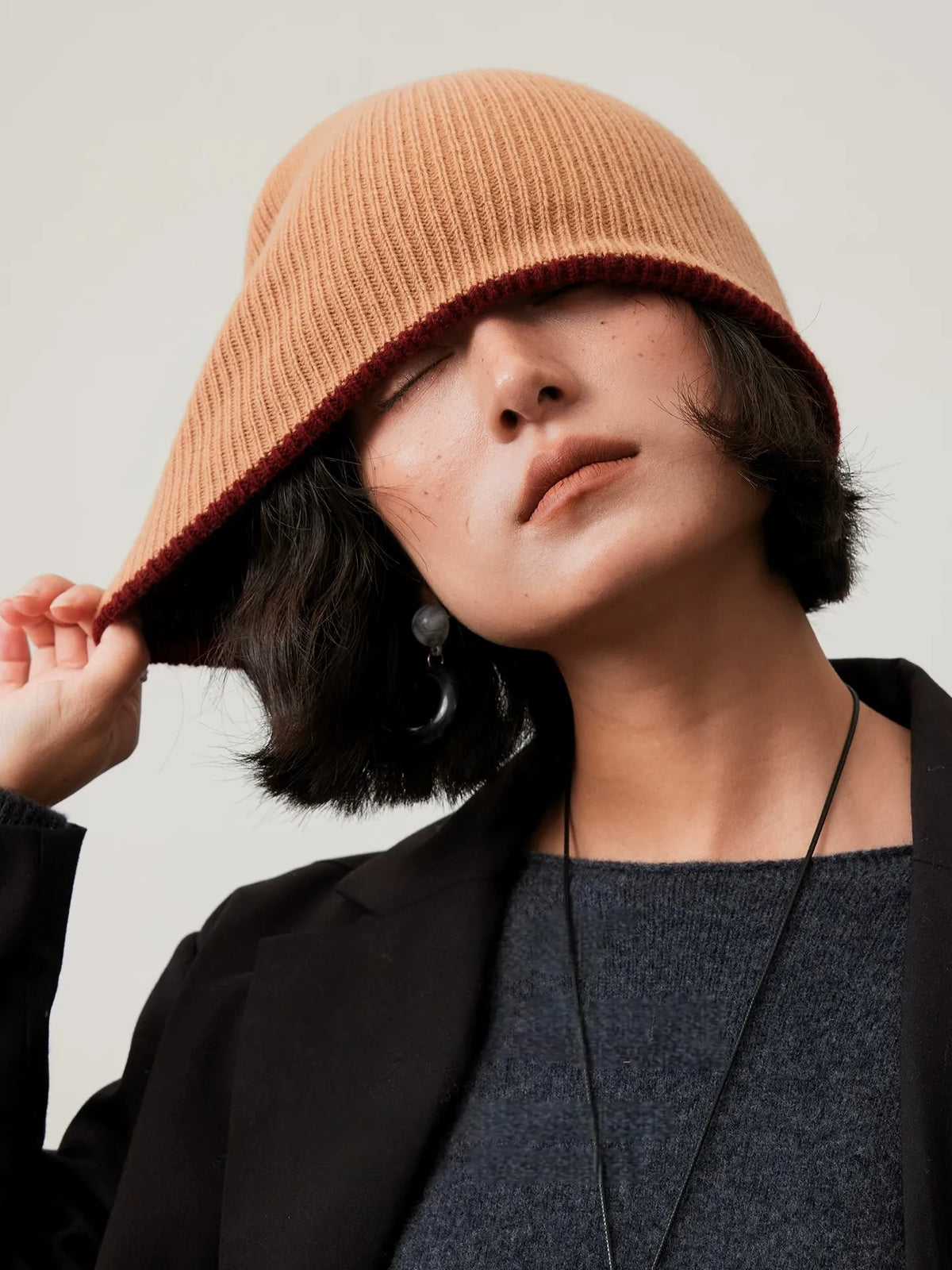 Model stretching the edge of a camel rib-knit wool beanie outward, showing its flexible construction.