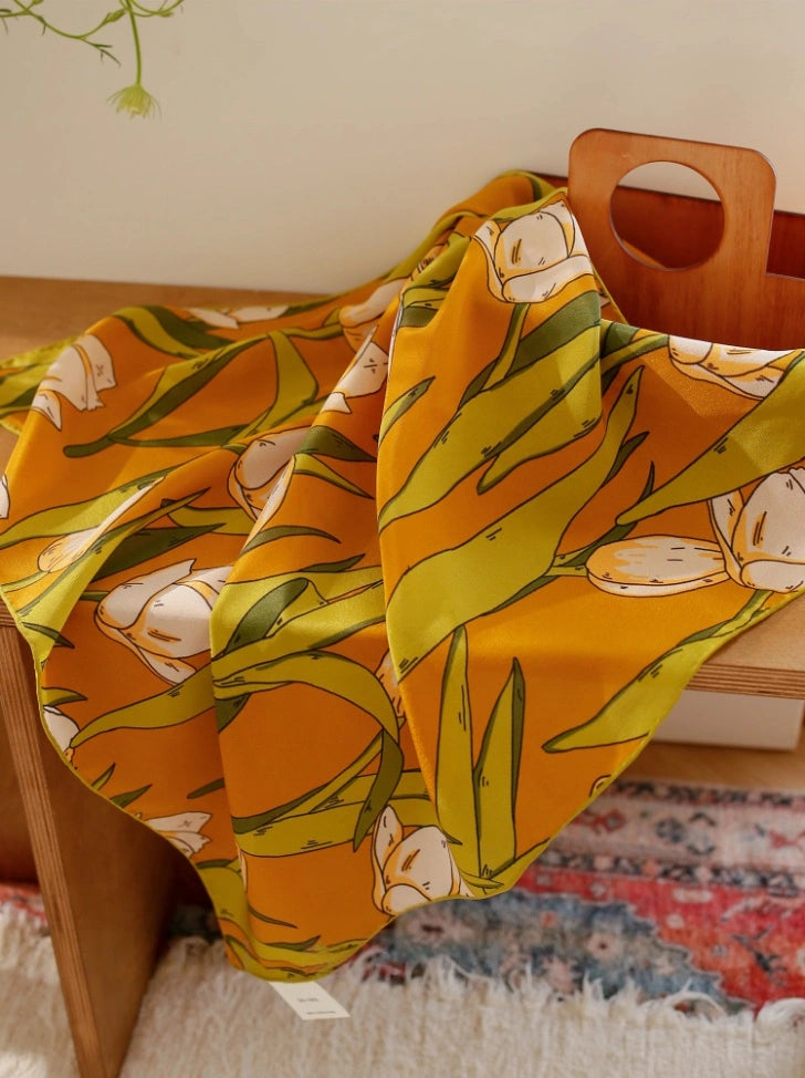 A yellow tulip floral printed silk square scarf made from 100% mulberry silk, laid flat on a tabletop for display.