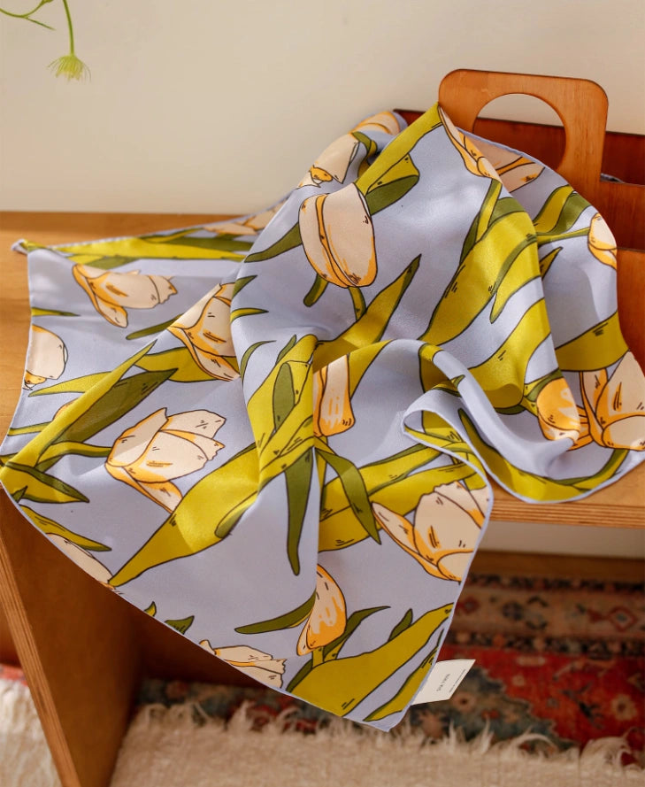 A blue tulip floral printed silk square scarf made from 100% mulberry silk, laid flat on a tabletop for display.