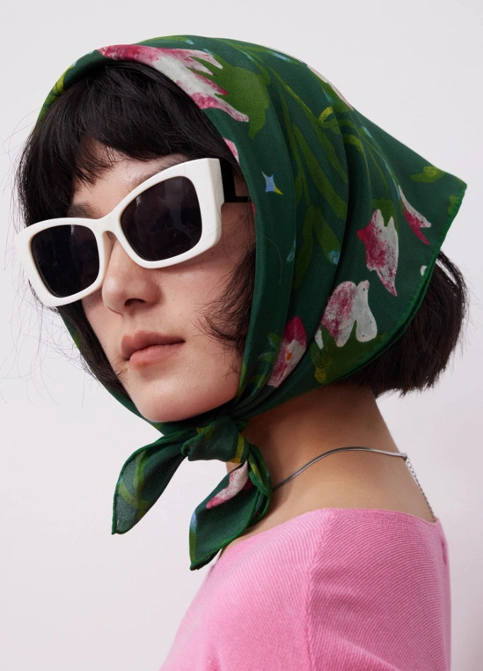 Woman wearing a Wilderness Green silk square scarf (63x63cm) as a headscarf, showcasing its floral mulberry silk design over a pink top, with a silver necklace.