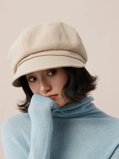 Close-up of a model wearing a beige newsboy cap with a blue sweater sleeve raised near her face.
