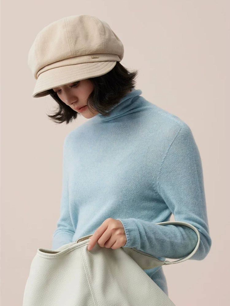 Model wearing a beige newsboy cap and light blue turtleneck while holding a pale green bag.