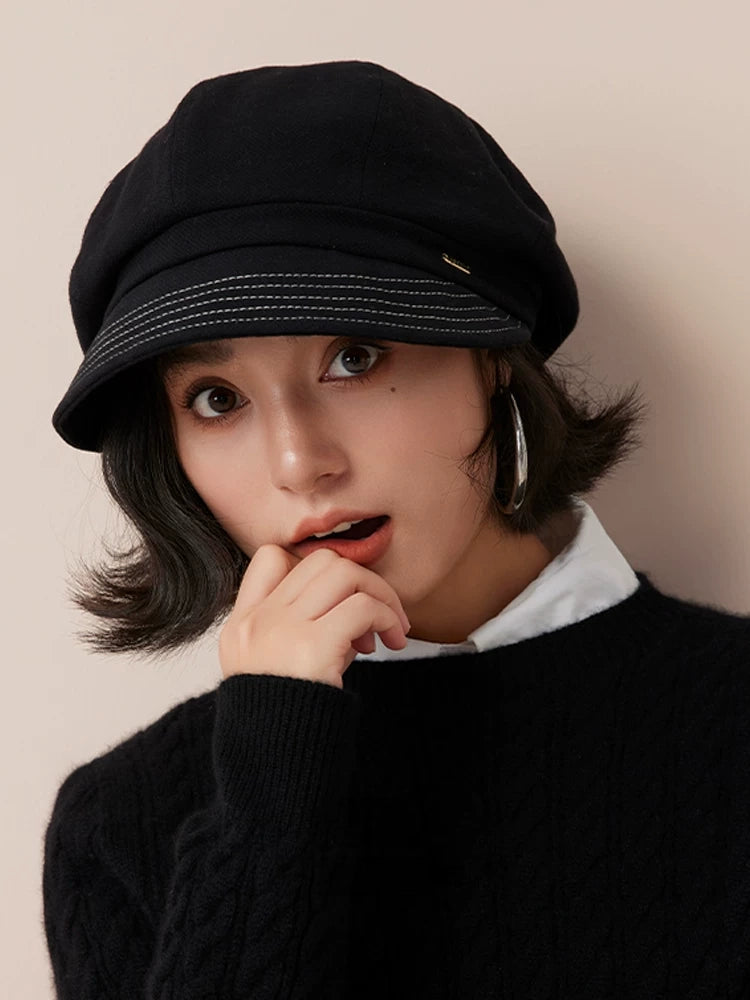 Front view of a black newsboy cap with a stitched brim, worn by a model resting her hand near her chin.