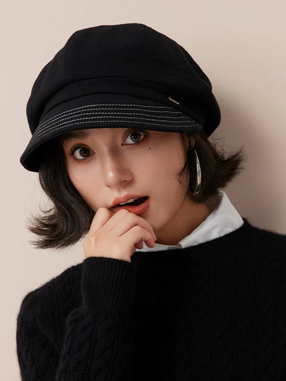Front view of a black newsboy cap with a stitched brim, worn by a model resting her hand near her chin.