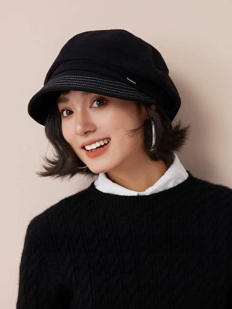Smiling model wearing a black newsboy cap with a black sweater and white shirt collar.