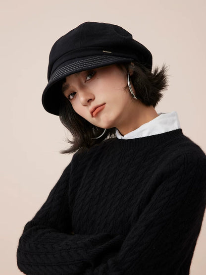 Model wearing a black newsboy cap with her head tilted and arms folded over a black sweater.