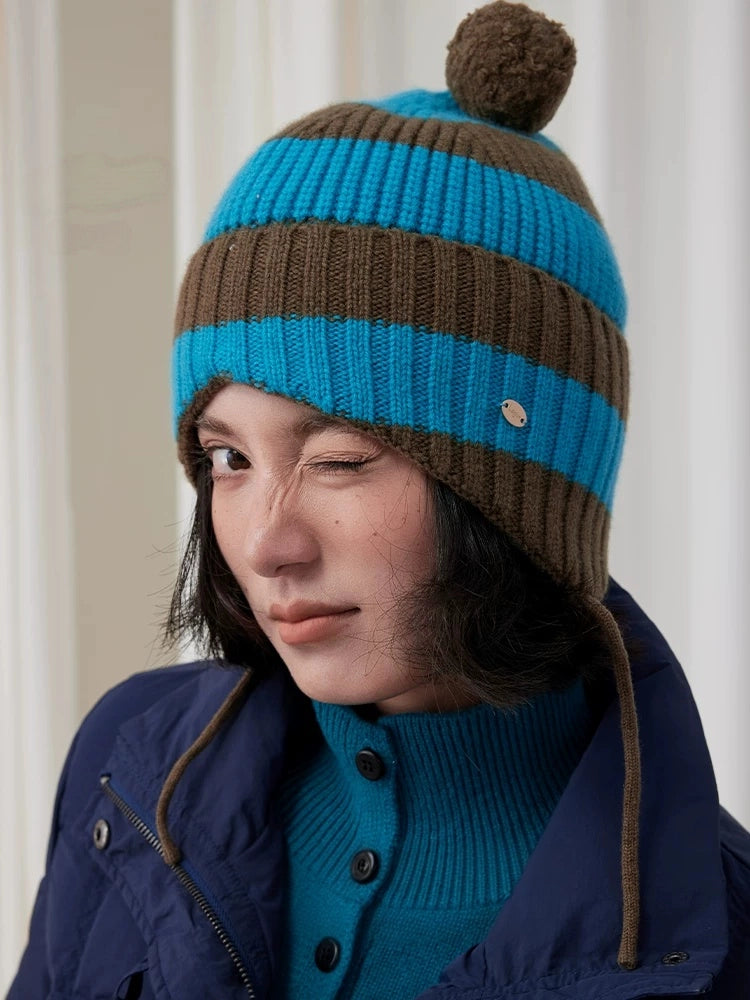 Front close-up of a model wearing a blue and brown striped wool earflap beanie with a brown pom pom.