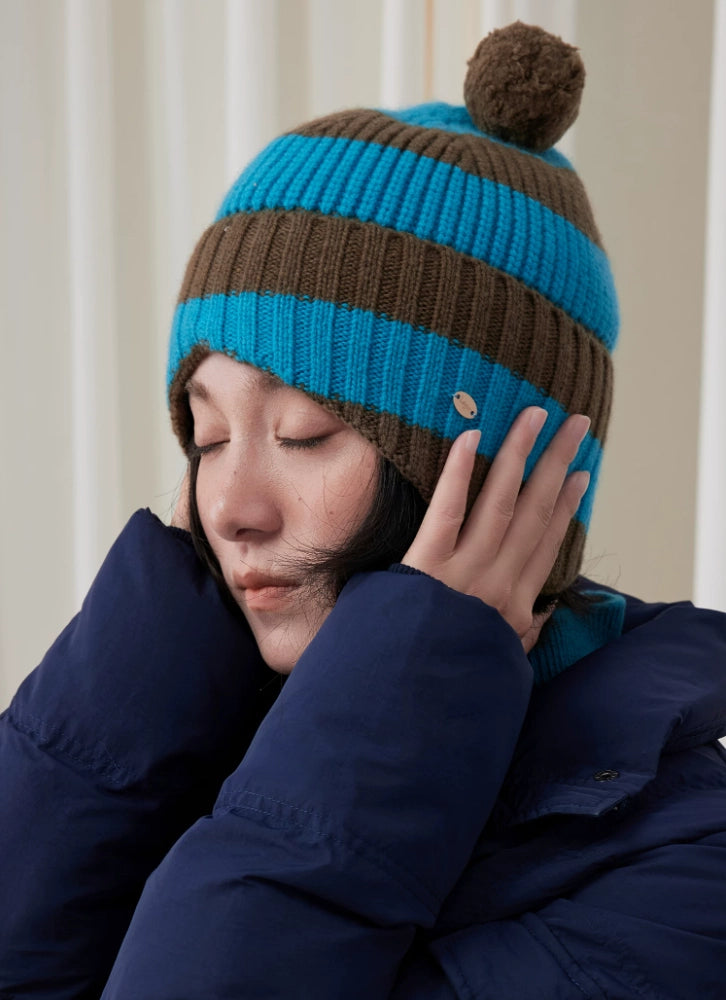 Model wearing a blue and brown striped wool earflap beanie, with both hands resting over the ears.