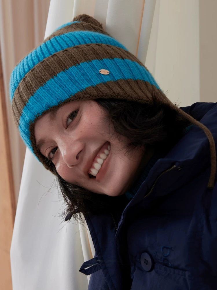 Tilted-head close-up of a model wearing a blue and brown striped wool earflap beanie.