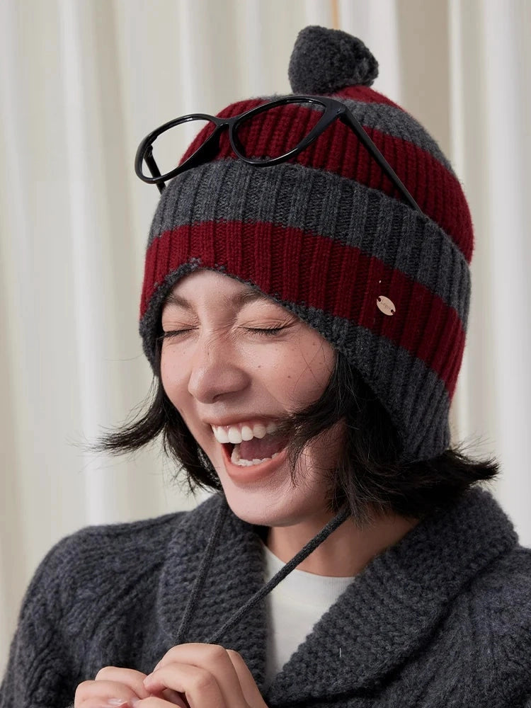 Model holding the grey ties of a burgundy and charcoal grey striped wool earflap beanie while laughing.