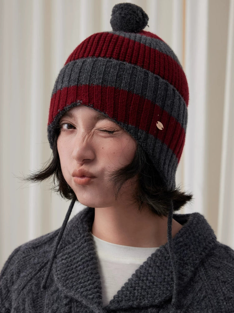Winking model wearing a burgundy and charcoal grey striped wool earflap beanie with a grey pom pom.