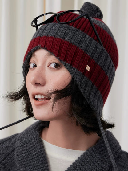 Close-up of a burgundy and charcoal grey striped wool earflap beanie with black glasses resting on top.