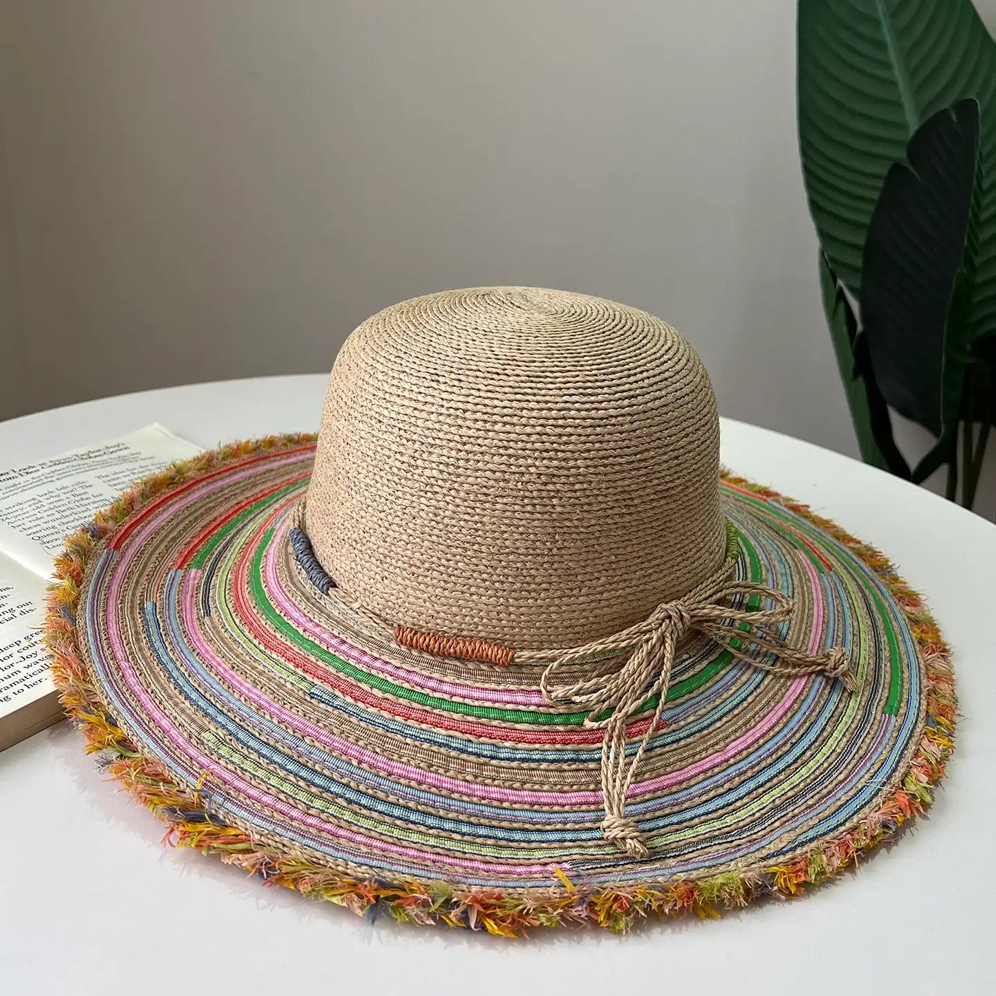 Beige raffia wide brim sun hat with colorful striped brim and frayed edge on white table.
