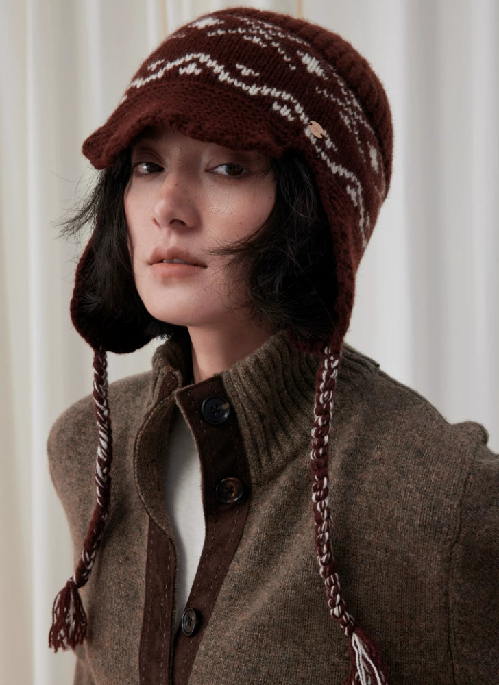 Front portrait of a model wearing a coffee brown Fair Isle earflap beanie with cream jacquard, a visor and braided ties.