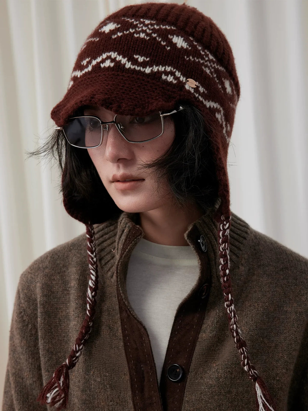 Model in sunglasses wearing a coffee brown Fair Isle earflap beanie with cream jacquard and braided ties.
