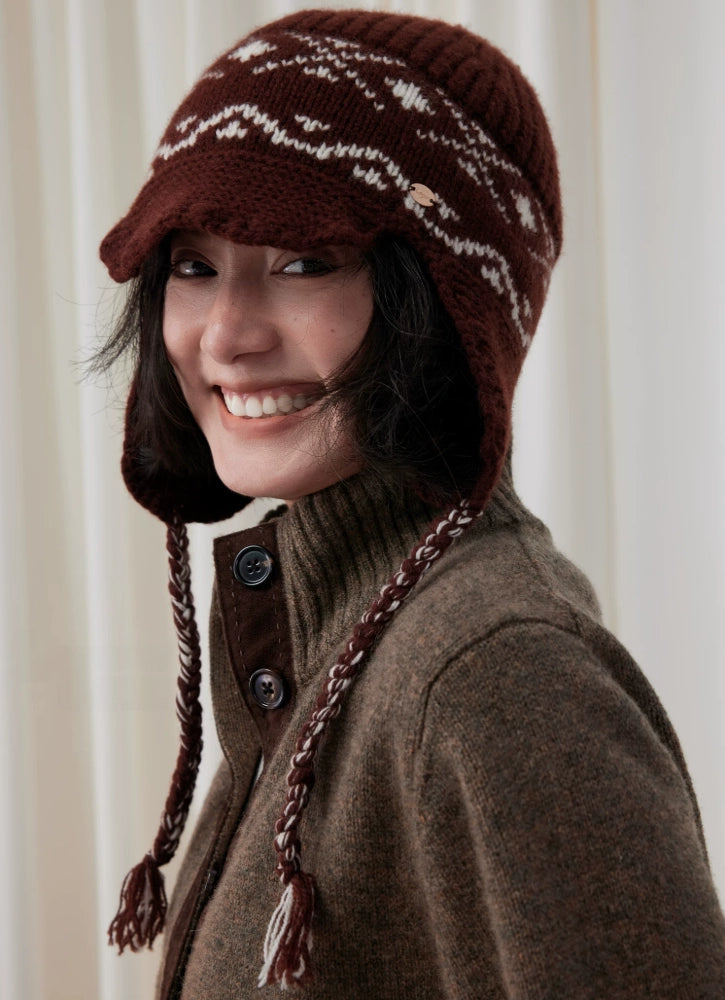 Smiling model wearing a coffee brown Fair Isle wool earflap beanie with a short visor and long braided ties.