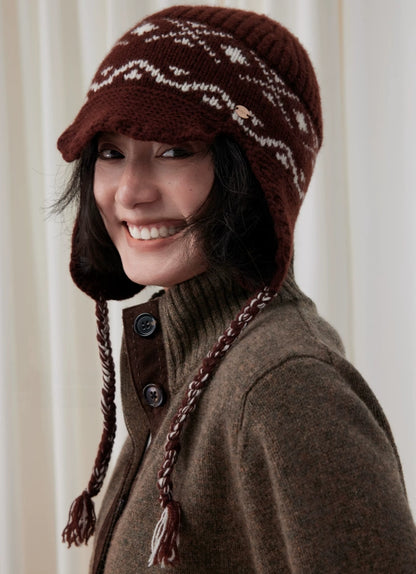 Smiling model wearing a coffee brown Fair Isle wool earflap beanie with a short visor and long braided ties.