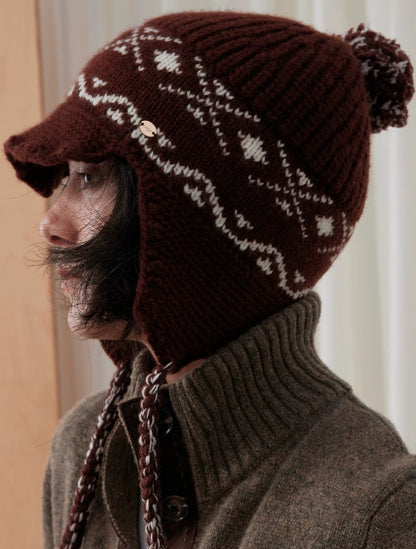 Side view of a coffee brown Fair Isle earflap beanie showing the short visor, braided tie and pom pom.