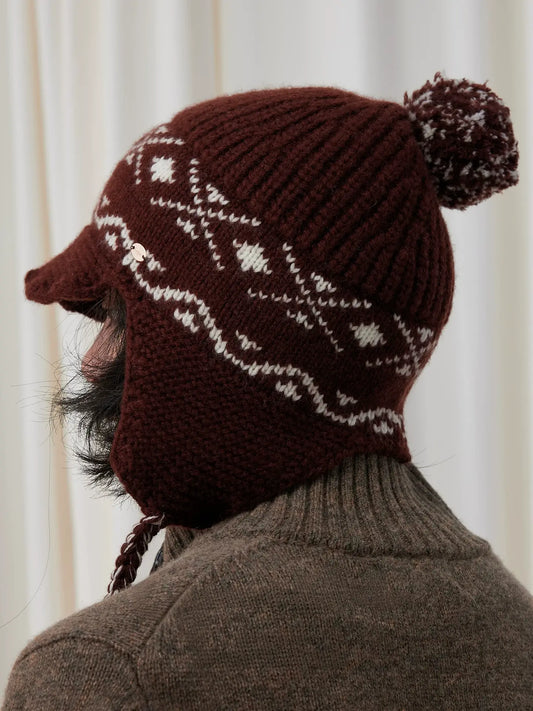 Rear three-quarter view of a coffee brown Fair Isle earflap beanie showing cream jacquard, a ribbed crown and pom pom.