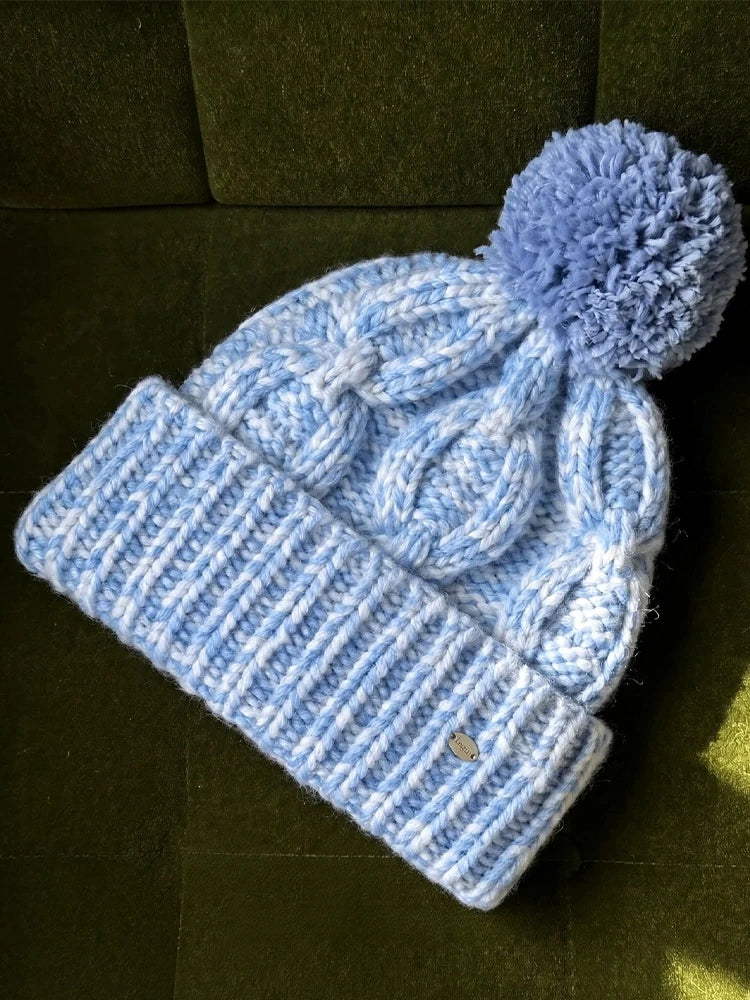 Light blue cable knit wool beanie with a matching blue pom pom and wide ribbed cuff, laid flat.