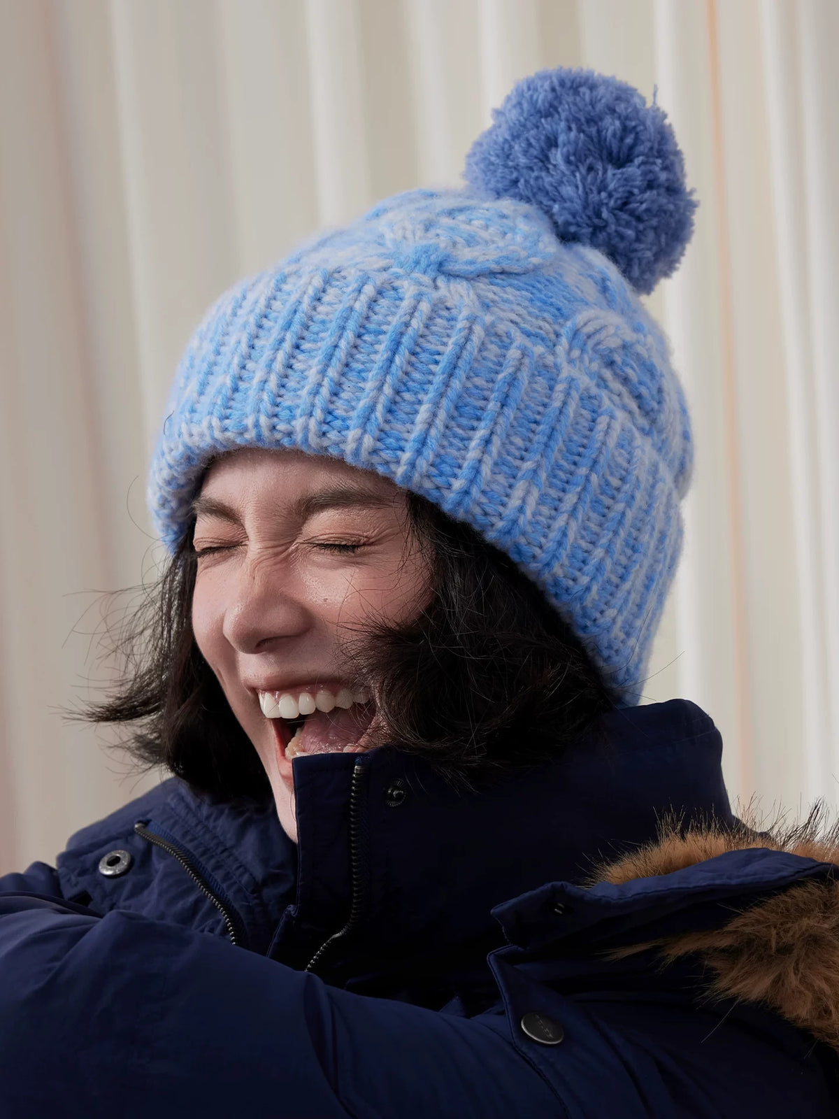 Model laughing in a light blue cable knit pom pom beanie with a wide ribbed cuff.