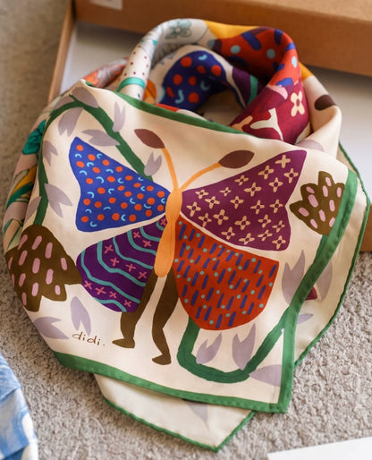 Folded cream mulberry silk square scarf with a burgundy, blue, and orange butterfly print and green border