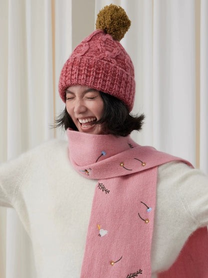 Model wearing a dusty pink cable knit beanie with a contrasting brown pom pom and pink scarf.