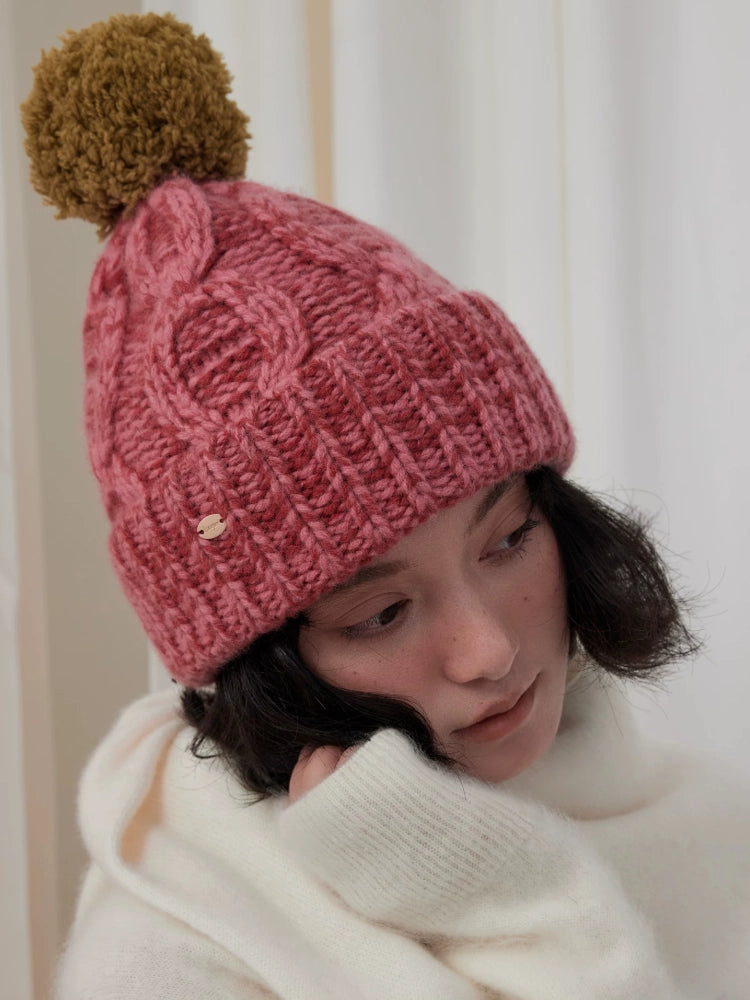 Angled close-up of a dusty pink cable knit pom pom beanie worn with a cream scarf.