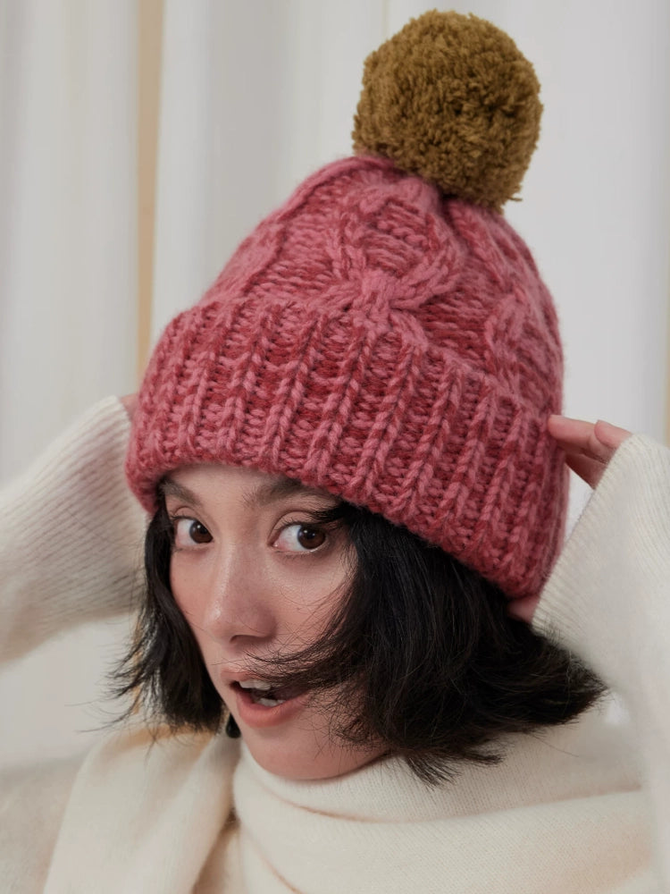 Front view of a model wearing a dusty pink cable knit beanie with a contrasting brown pom pom.
