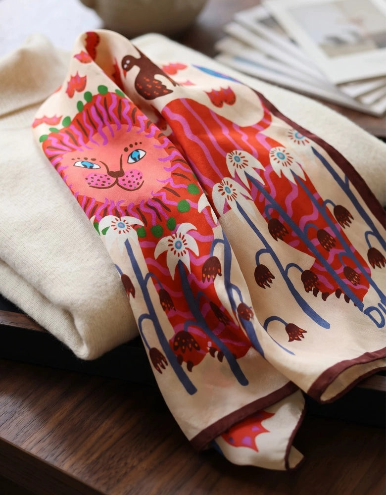 Close-up of folded mulberry silk scarf showing the pink lion, red flowers, and illustrated animal details