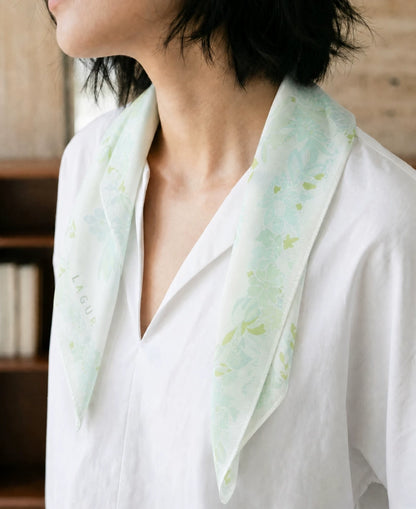 Woman wearing a tender green floral mulberry silk scarf loosely draped over a white blouse