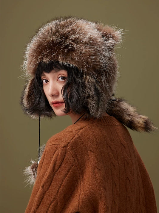 Model wearing a brown-grey fur trapper hat in three-quarter view, showing long ear flaps and a raccoon-style tail.