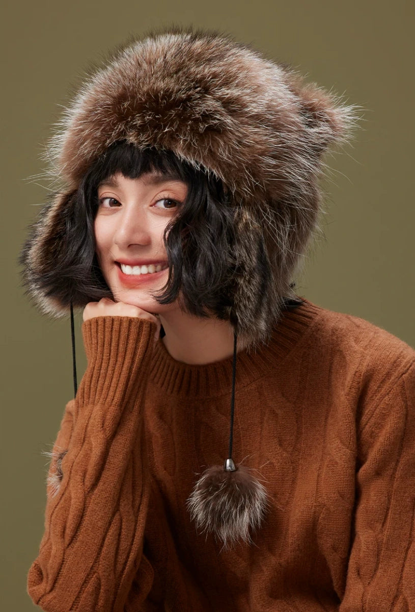 Front view of a model wearing a brown-grey fur trapper hat with plush ear flaps and hanging pom-pom cords.