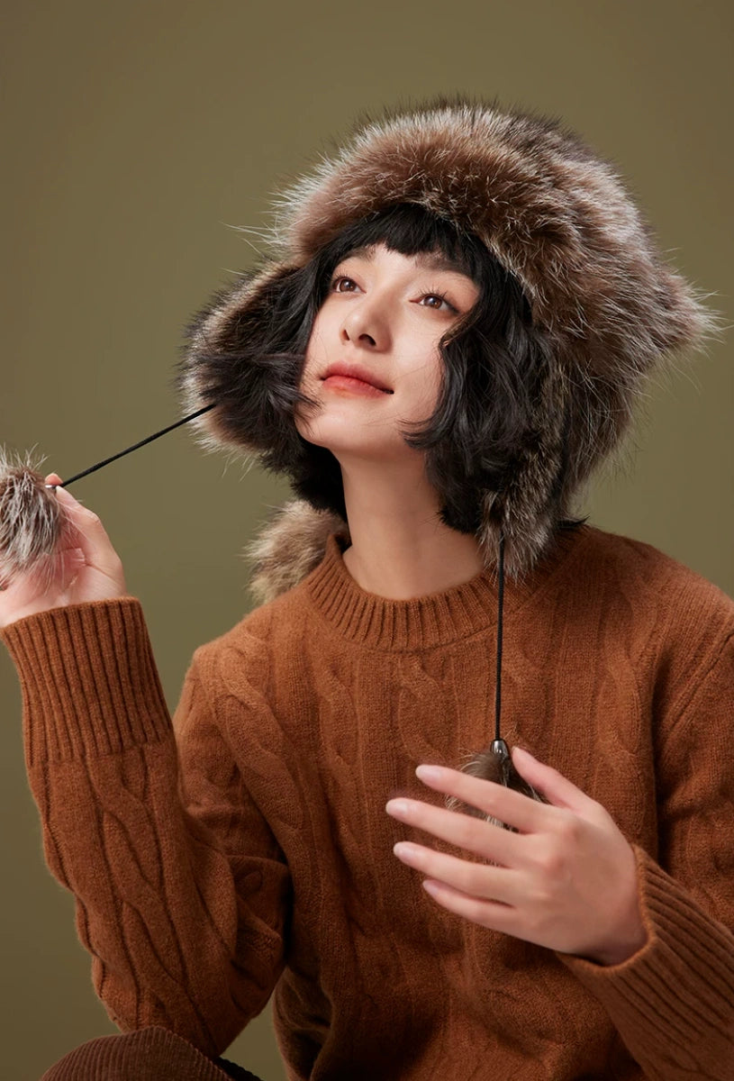 Model wearing a brown-grey fur trapper hat while holding a pom-pom cord, highlighting the plush ear flaps.