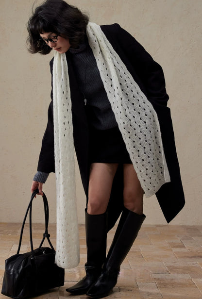 Model wearing a cream white wave pointelle cashmere scarf draped open over a black coat with knee-high boots.