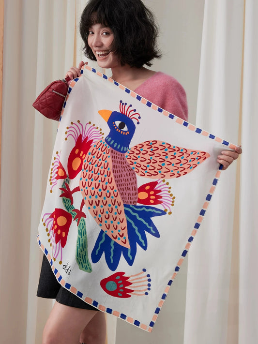 Woman holding an unfolded ivory silk square scarf with a colorful blue and coral bird print
