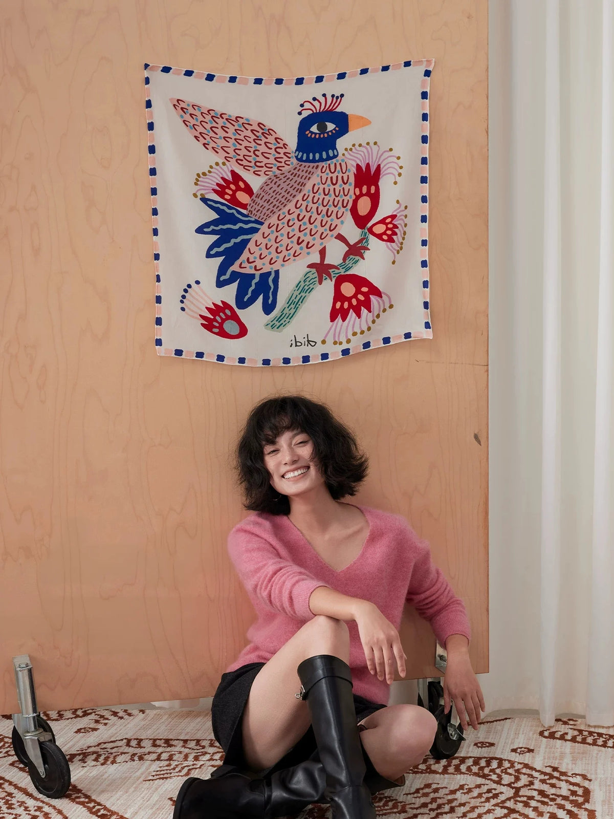 Illustrated bird mulberry silk scarf displayed as wall art above a woman seated in a pink sweater