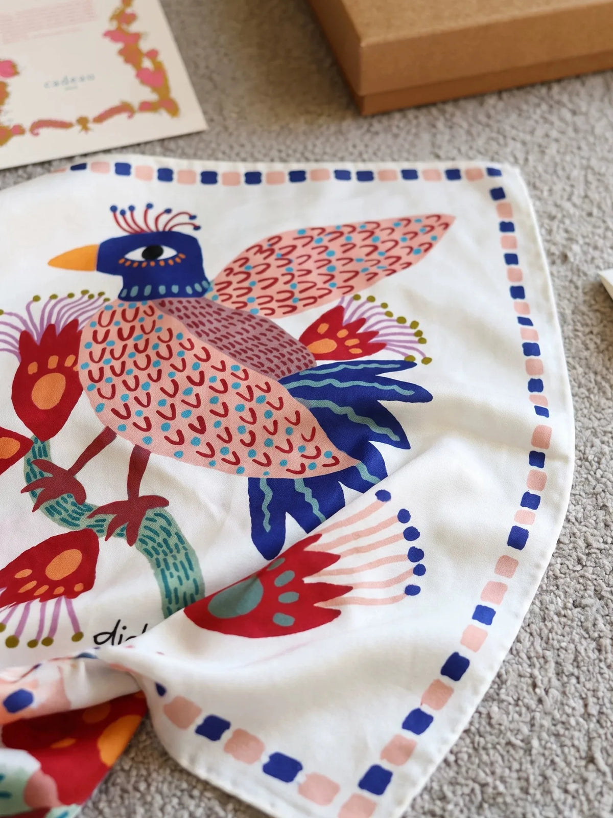 Close-up of an ivory mulberry silk square scarf featuring a blue, coral, and green folk bird design
