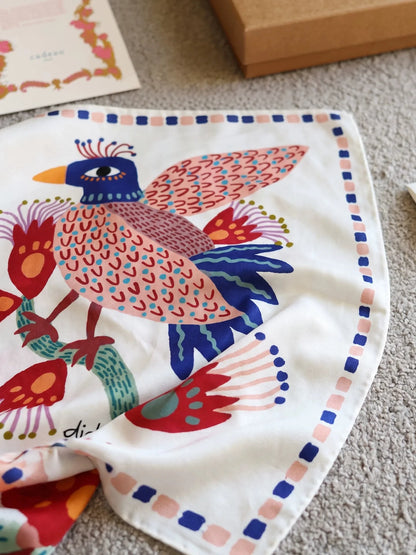Close-up of an ivory mulberry silk square scarf featuring a blue, coral, and green folk bird design