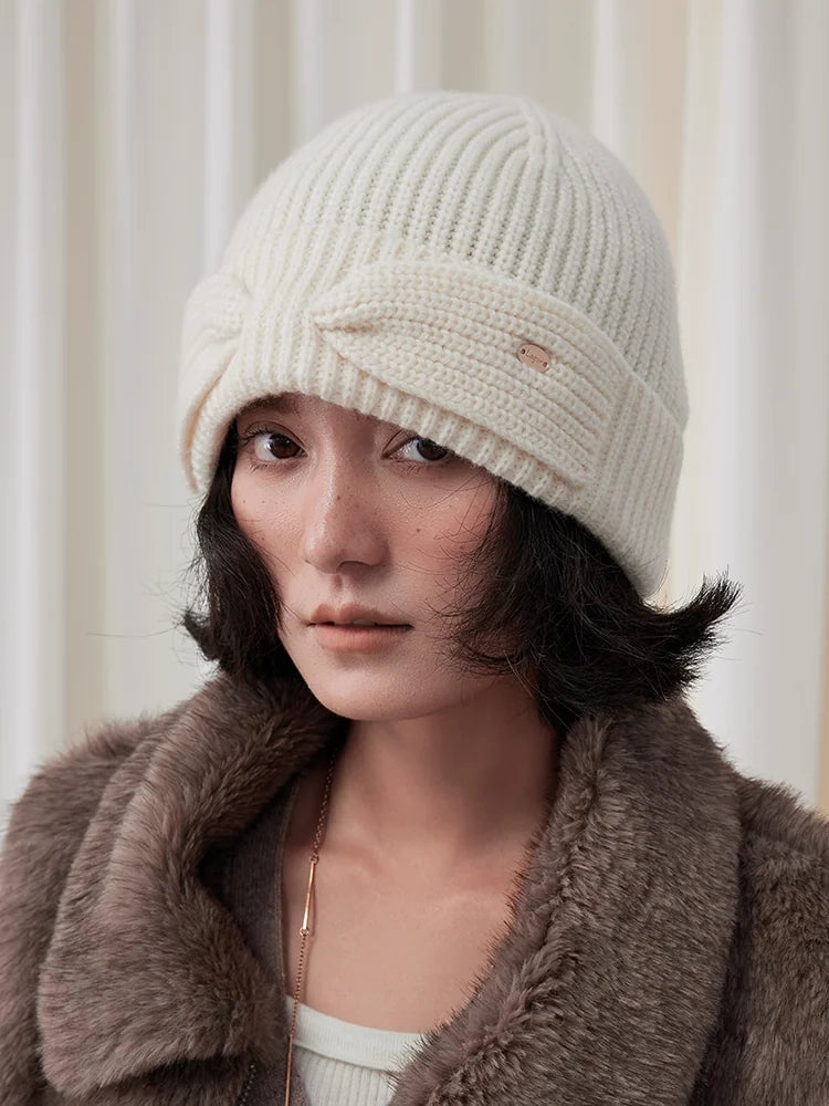 Model wearing a white wool bow beanie with vertical ribbing, a folded cuff, and a small gold-toned accent.