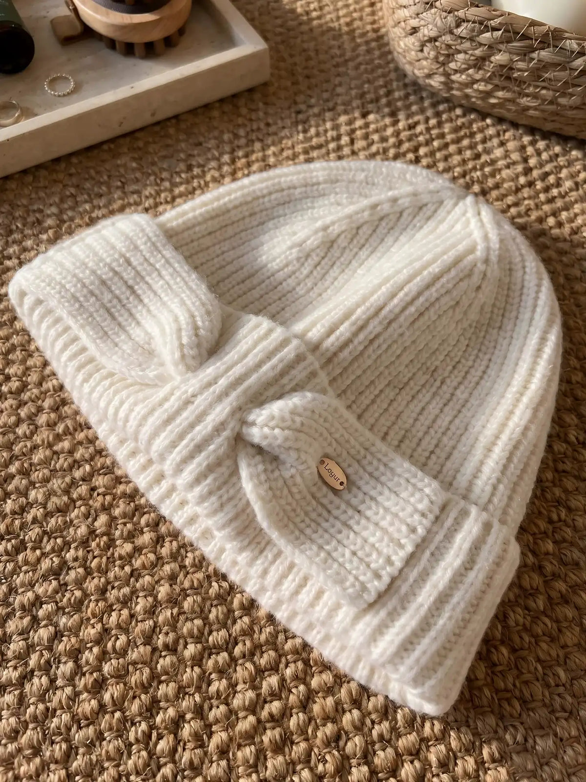 White wool bow beanie laid flat on a woven mat, showing the ribbed knit, folded cuff, and bow detail.