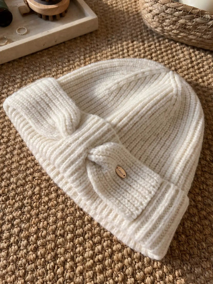 White wool bow beanie laid flat on a woven mat, showing the ribbed knit, folded cuff, and bow detail.