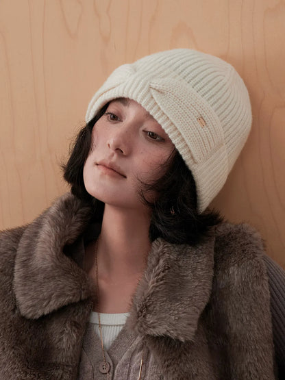 Model tilting her head in a white wool bow beanie, with the folded cuff and bow visible against a wooden wall.