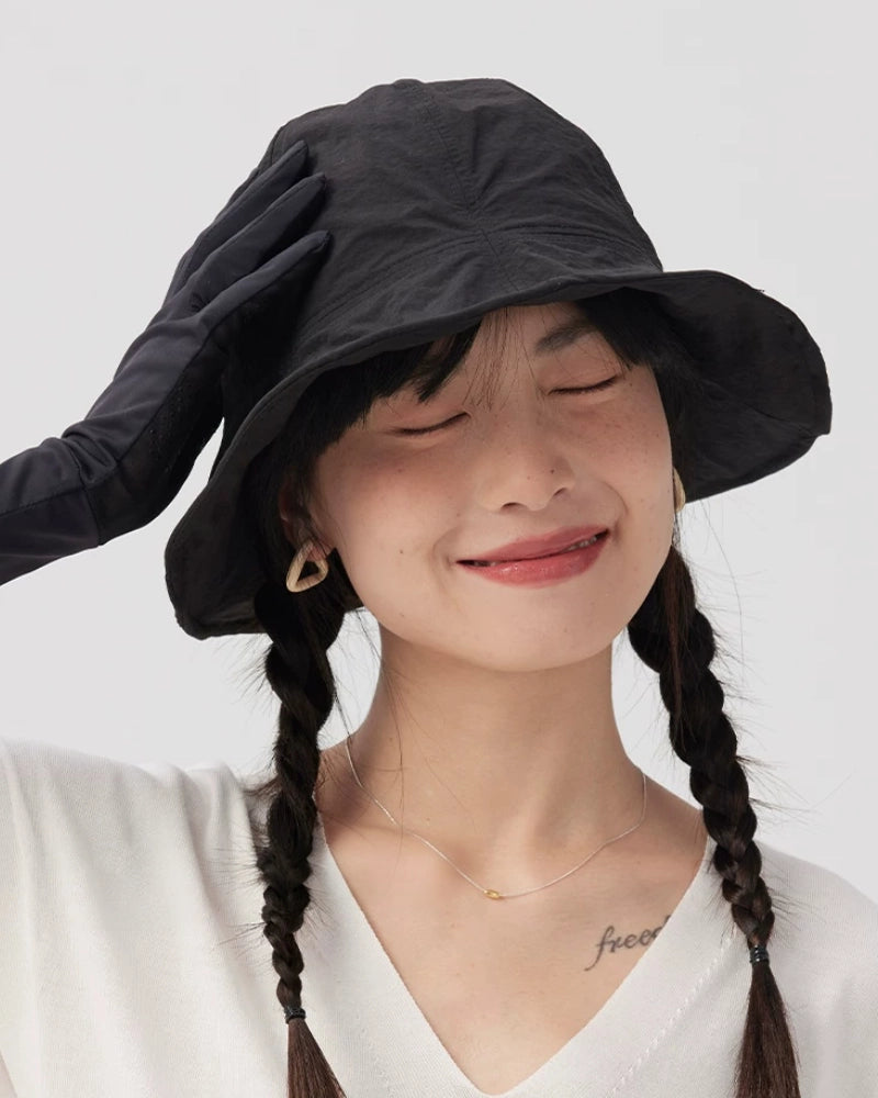 Woman smiling in a black wide brim bucket hat with one gloved hand resting on the crown.