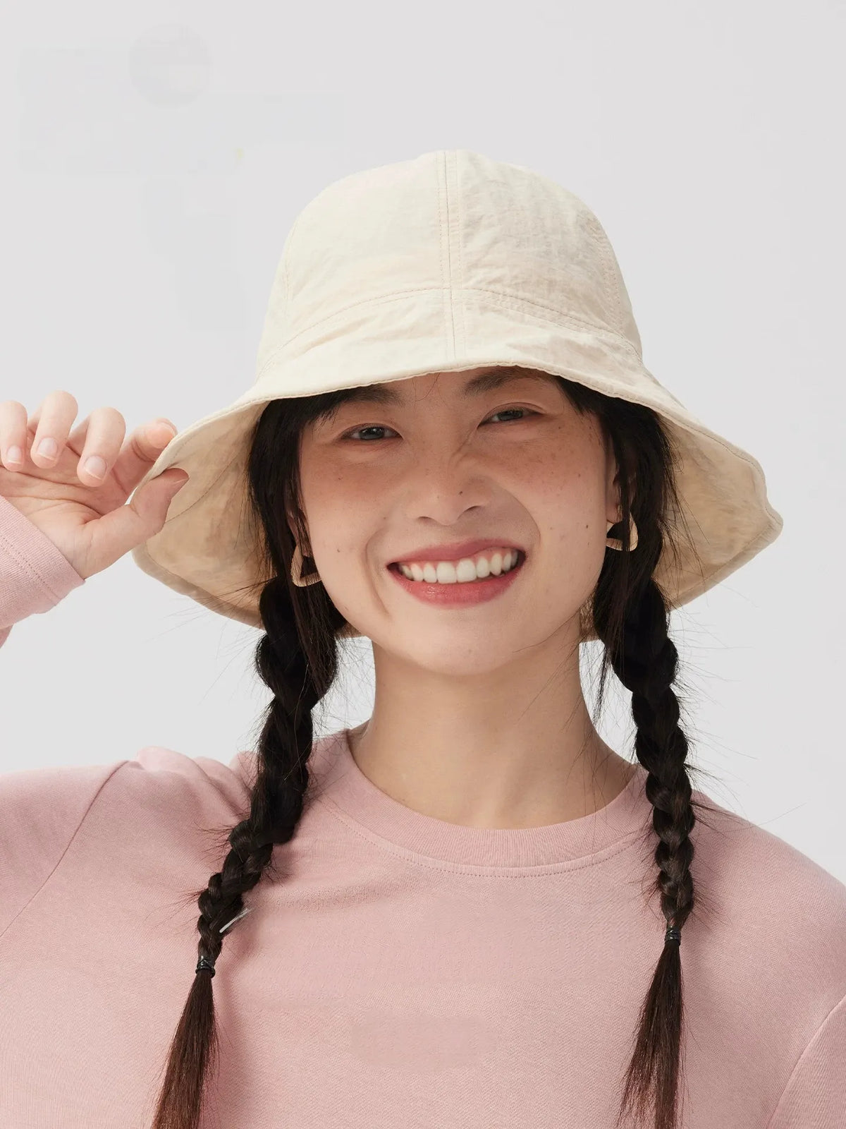 Woman smiling and holding the brim of an ivory bucket hat, styled with twin braids and a pink top.