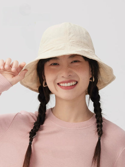 Woman smiling and holding the brim of an ivory bucket hat, styled with twin braids and a pink top.